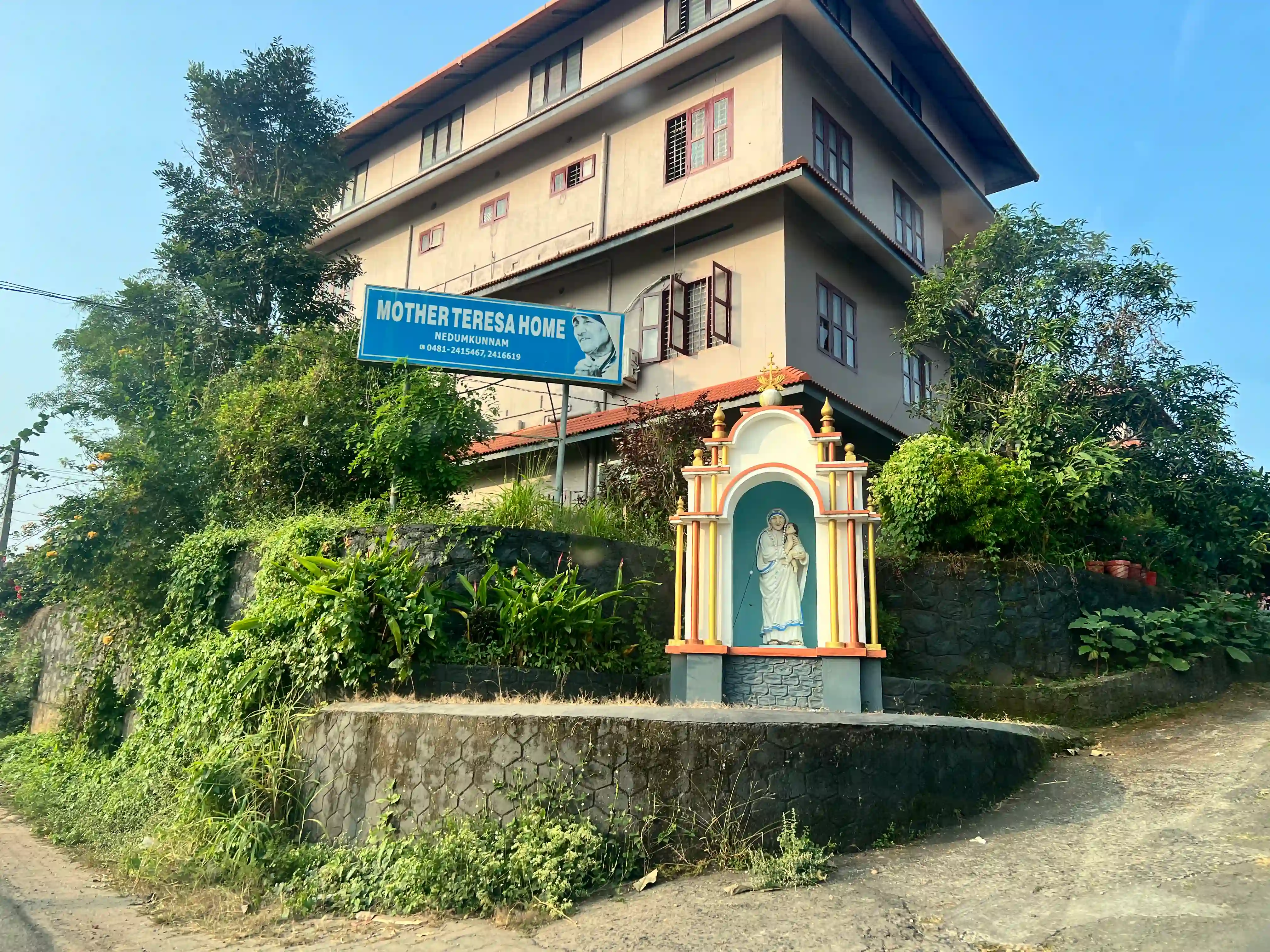 The roadside signboard marking the turn to Mother Teresa Home, Nedumkunnam, beside a small shrine to Our Lady.